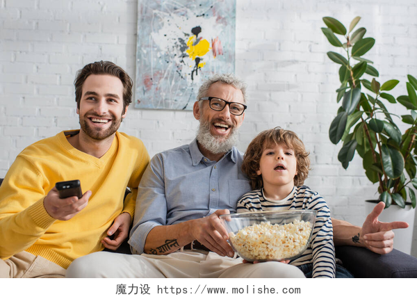 在客厅看电视的祖孙三代在家里用爆米花笑着男人和男孩看电视 
